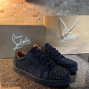 Christian Louboutin mörkblå sneakers mocka - Snygga mörkblå sneakers från Christian Louboutin i mocka med svarta nitar på tå och häl. Skorna har klassisk snörning, rund tå och den ikoniska röda sulan. Perfekta för dig som vill sticka ut med exklusiv streetstyle. Du får med allting som syns på bilderna. 