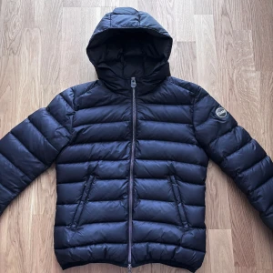 Svart Colmar jacka med luva - Säljer en svart pufferjacka från Colmar med luva och snygga horisontella quiltade partier. Jackan har dragkedja framtill, två fickor med dragkedja och Colmar-logga på ärmen. Perfekt för dig som vill ha en stilren och varm jacka med modern look. Använd i 2 vintrar. På lappen står det storlek 42 men den passar mig som brukar ha XS/S. Jackan är i bra skick i övrigt, men tyget har gått upp lite i ena ärmen och hängöglan har lossnat på ena sidan (går enkelt att fixa om man vill, se bild) 💓