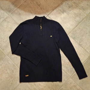 Svart stickad tröja från Aiopeson - Svart stickad tröja från Aiopeson med ribbat mönster och half zip-dragkedja i guld. Tröjan har hög krage och långa ärmar, samt en liten metallbricka med logga på bröstet. Perfekt för lager-på-lager under kyliga dagar.