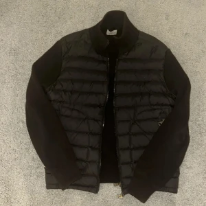 Moncler cardigan  - Säljer en svart dunjacka från Moncler med quiltad front och ärmar i stickad ull. Jackan har dragkedja framtill och en clean, stilren look. Perfekt för dig som vill ha en snygg och exklusiv jacka med både värme och streetkänsla.