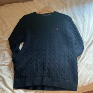 Mörkblå kabelstickad tröja Polo Ralph Lauren - Kabelstickad mörkblå tröja från Polo Ralph Lauren med klassisk rund hals och röd broderad logga på bröstet. Tröjan är långärmad och har ribbade muddar vid ärmslut och nederkant. Perfekt för dig som gillar preppy och stilren look. Har aldrig använt 