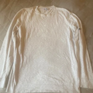 Beige långärmad tröja från Stone Island - Snygg beige långärmad tröja från Stone Island med rund hals och två svarta knappar på ärmen. Tröjan är i mjuk bomullsblandning och har en clean, stilren look som passar perfekt till jeans eller chinos.