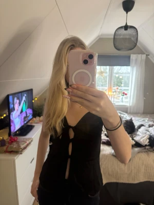 Svart omlott linnetopp med knyt - Säljer en svart ärmlös linnetopp med djup v-ringning och tre knyt framtill. Toppen har en croppad passform och är tillverkad i ett slätt, lätt material som ger en snygg siluett. Perfekt för dig som gillar trendiga och stilrena plagg.