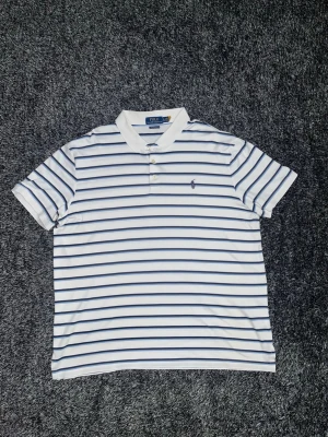 Randig pikétröja från Polo Ralph Lauren - Snygg vit pikétröja från Polo Ralph Lauren med marinblå och ljusblå ränder. Klassisk krage, tre knappar och broderad logga på bröstet. Modellen är custom slim fit och passar perfekt till en avslappnad men stilren look. Storlek XL men passar L väldigt väl också. En riktigt fin tröja som är perfekt nu inför lite ljusare tider. Skick 10/10. Tveka inte på höra av dig vid minsta intresse😄. /Isak 