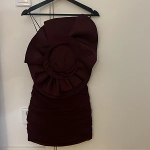 Vinröd kort klänning  - Säljer en vinröd kort klänning från Magda Butrym x H&M med en stor dekorativ blomdetalj på ena axeln och plisserade partier. Klänningen är oneshoulder och har en tight passform som framhäver siluetten. Perfekt för dig som vill sticka ut med en unik design.