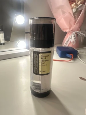 COSRX Snail Radiance Dual Essence - En genomskinlig pumpflaska med svart lock som innehåller COSRX Advanced Snail Radiance Dual Essence. Essencen är känd för sin återfuktande och lystergivande effekt, med snigelsekret som huvudingrediens. Flaskan rymmer 80 ml och har en stilren design.