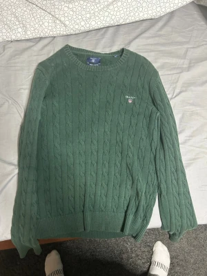 Grön kabelstickad tröja från GANT - Säljer en grön kabelstickad tröja från GANT i storlek S. Tröjan har rund hals, ribbade muddar och GANT-logga broderad på bröstet. Perfekt för dig som gillar klassisk stil med snygga detaljer och mjukt material. Passar även M
