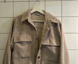 Beige manchester overshirt med fickor - Säljer en beige overshirt i manchester med bred krage och stora bröstfickor. Jackan har knäppning med knappar framtill och ett avslappnat snitt. Perfekt för lager-på-lager och chill stil.