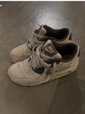 Nike Air Max sneakers i beige mocka - Nike Air Max sneakers med beige mocka och svarta detaljer. Skorna har guldfärgad Nike-logga på tungan och sidan, samt synlig Air Max-sula med brun yttersula. Klassisk snörning och rund tå. Perfekt för dig som gillar retrostil och streetwear. Kostar att köpa nya ca 3000 nu