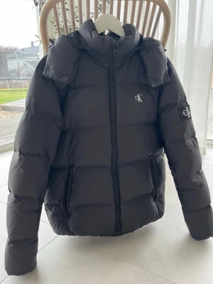 Svart pufferjacka från Calvin Klein - Jackan är i väldigt bra sick och använd 1 och en halv säsong. Den har ett pyttelitet hål på luvan, men syns inte när den används. Det finns ett litet hål i fickan intill ytterkanten på fickan men märks inte att det är ett hål där. Strl:M  Herr Hör av er vid fler frågor.