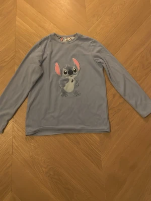 Disney Stitch pyjamas tröja blå - Mjuk och mysig blå långärmad fleece tröja från Disney med stort Stitch-tryck framtill. Tröjan har rund hals och är perfekt för dig som gillar Lilo & Stitch. Passar dig som vill ha något bekvämt och snyggt med karaktärsmotiv.