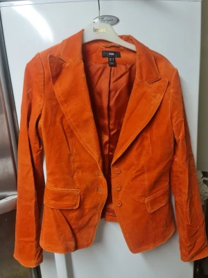Orange manchesterkavaj från H&M - Snygg orange kavaj från H&M i mjuk manchester. Klassisk modell med krage, tre knappar framtill och två fickor med lock. Perfekt för dig som vill sticka ut med färg och cool retrovibe. Långärmad och figurnära passform.