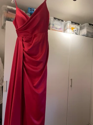 Röd långklänning med drapering - Säljer en elegant mörk rosa långklänning med smala axelband och v-ringad omlottliv. Klänningen har snygg drapering längs sidan och är gjord i ett glansigt, mjukt material som ger en lyxig känsla. Perfekt för dig som vill sticka ut med en färgstark look.