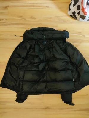 Svart pufferjacka från Polo Ralph Lauren - Svart pufferjacka från Polo Ralph Lauren med huva och dragkedja framtill. Jackan har puffad design, två sidofickor med dragkedja och Polo-logga i rött på bröstet. Perfekt för kalla dagar och har en riktigt schysst streetstil.