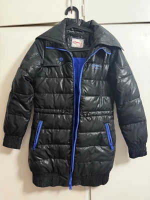 Svart pufferjacka från O'Neill - Svart pufferjacka från O'Neill med blå detaljer och dragkedjor. Jackan har hög krage, långa ärmar med resår och två fickor med blå dragkedja. Materialet är glansigt syntet och jackan har quiltad design samt snörning i midjan. Perfekt för kalla dagar.