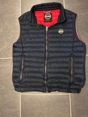 Mörkblå dunväst från Colmar - Snygg mörkblå dunväst från Colmar med quiltad design och röd insida. Västen har två fickor med dragkedja och en hög krage. Materialet är polyester och västen stängs med en rejäl dragkedja framtill. Perfekt för lager-på-lager under kyliga dagar.