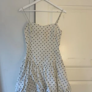 Vit prickig kort klänning Bershka - Supersöt vit kort klänning från Bershka med svarta prickar och tunna axelband. Klänningen har en figurnära överdel och volangig kjol, perfekt för sommarens alla tillfällen. Tillverkad i bomull för en skön och luftig känsla.