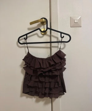 Brun volang linnetopp från Zara - Säljer en brun linnetopp från Zara i storlek S. Toppen har tunna axelband och flera lager med volanger framtill, medan baksidan är ribbad och slät. Perfekt för dig som gillar detaljer och vill ha en unik topp i garderoben.