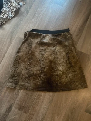 Brun kortkjol med ormmönster från Figa Str XS - Säljer en brun kortkjol från Figa med coolt ormmönster och lätt glansig yta. Kjolen har en svart midjeresår och är super snygg💕