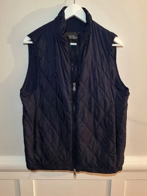 Väst från Oscar Jacobson - Quiltad mörkblå dunväst från Oscar Jacobson med hög krage och dragkedja framtill. Västen har ett stilrent rutmönster och är tillverkad i syntetmaterial. Perfekt inför våren, mycket grisch! Nästan aldrig använd, köptes förra våren