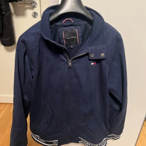 Mörkblå overshirt från Tommy Hilfiger - Snygg mörkblå overshirt från Tommy Hilfiger med dragkedja, ståkrage och tryckknappar. Jackan har två fickor framtill och logoband med Tommy Hilfiger-tryck längs nederkanten. Perfekt för dig som gillar stilren design och klassiska detaljer. Storlek S men passar även M, jackan är använd en gång endast och har inga defekter eller sådant!