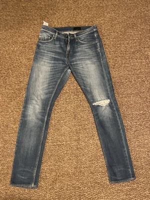 Blå skinny jeans med slitning - Säljer ett par blå skinny jeans med snygg slitning på höger knä och tvättade detaljer. Klassisk femficksmodell med normal midja och smal passform. Perfekta för dig som gillar en trendig och avslappnad stil.