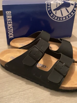Svarta sandaler från Birkenstock  - Säljer ett par klassiska svarta Birkenstock sandaler med två breda remmar och justerbara spännen. Fotbädden är mjuk och ergonomisk med kork och mocka, yttersulan är svart och mönstrad för extra grepp. Perfekt för chill dagar och enkel stil.