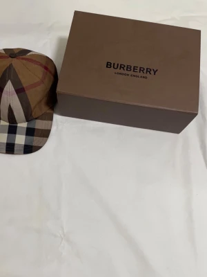 Rutig Burberry keps i bomull - Säljer en snygg Burberry keps i klassiskt rutigt mönster med bruna, beige, svarta och vita toner. Kepsen är tillverkad i 100% bomull och har justerbar rem med metallspänne där Burberry är ingraverat. Perfekt för dig som vill ha en ikonisk accessoar.