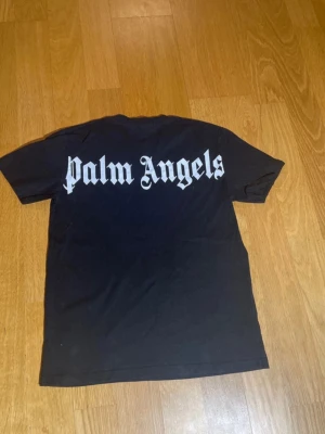 Svart Palm Angels t-shirt med tryck - Svart t-shirt från Palm Angels i bomull med stort vitt tryck på ryggen och mindre logga vid halsen. Klassisk rund hals och korta ärmar. Perfekt för dig som gillar streetwear och vill sticka ut med en fet logga på ryggen.