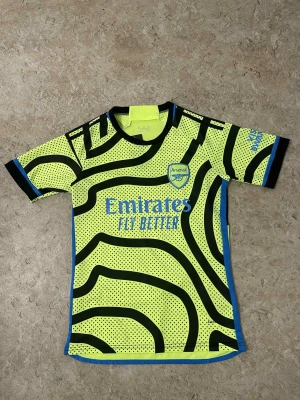 Arsenal fotbollströja gul/svart - Cool Arsenal fotbollströja i neon-gult med svarta och blå detaljer. Tröjan har ett unikt randigt mönster med prickar och klubbens logga på bröstet. Materialet är lätt polyester som andas, perfekt för fotbollsträning eller match.