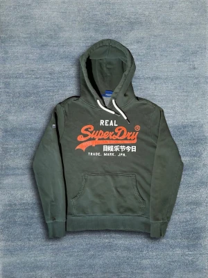 Superdry Hoodie - Grön hoodie ifrån Superdry. Tröjan är i bra skick men har några bristningar i den oranga texten. Annars är tröjan i väldigt bra skick. Hör av dig vid frågor eller fler bilder!🙌 