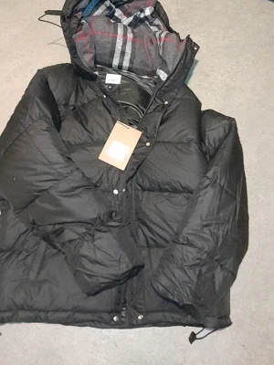 Burberry Jacka - [Blir till väst] Svart pufferjacka från Burberry med huva och rutig insida i grått, vitt och rött. Jackan har dragkedja och knappar framtill, samt snörning vid nederkanten. Märkespatch på ärmen och klassisk Burberry-rutig lining. Perfekt för kalla dagar.