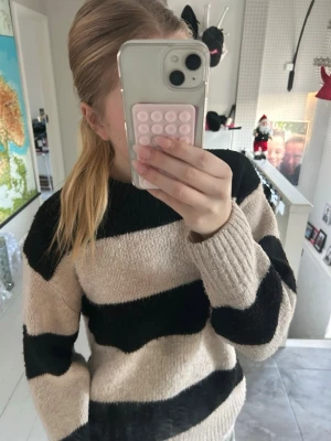 Beige och svart randig stickad tröja - Mysig stickad tröja med breda svarta och beige ränder. Tröjan har en klassisk rund halsringning och långa ärmar. Perfekt för att hålla sig varm under kyliga dagar och passar till både jeans och kjol.