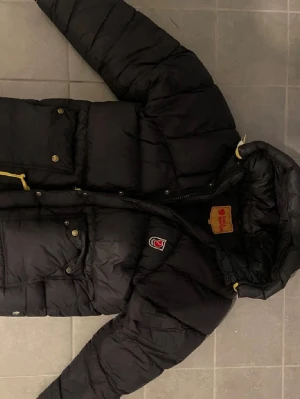 Fjällräven Jacka Dam XXS - Svart dunjacka från Fjällräven med gul snörning i huvan och klassisk logga på bröstet. Jackan har flera fickor med knappar och dragkedja framtill. Perfekt för kalla dagar och har en schysst, puffig look.
