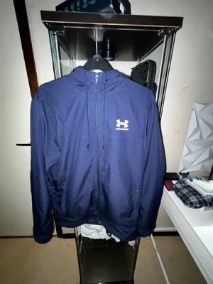 Blå vindjacka från Under Armour - Snygg marinblå vindjacka från Under Armour med huva och dragkedja framtill. Jackan har meshfoder på insidan, snörning vid huvan och logga på bröstet. Perfekt för aktiva dagar och träning utomhus. Trasig dragkedja 