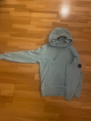 Ljusblå hoodie från C.P. Company - Säljer en ljusblå hoodie från C.P. Company med klassisk linsdetalj på ärmen och magficka. Tröjan har dragsko i huvan och ribbade muddar. Perfekt för dig som gillar streetwear och snygga detaljer.
