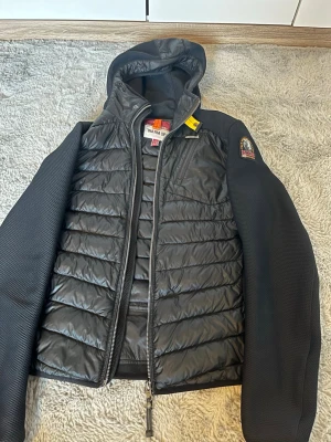 Svart cardigan från Parajumpers - Svart cardigan från Parajumpers med huva och quiltad framsida. Jackan har släta, svarta ärmar i syntetmaterial och dragkedja framtill. Märkespatch på ena ärmen och praktiska fickor med dragkedja. Perfekt för dig som gillar stilren och sportig look.