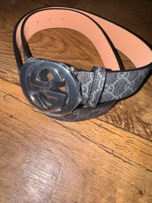 G bälte - Säljer ett bälte för ett bra pris. Bältet är svart med grått monogrammönster och har en bred, klassisk form. Insidan är beige och bältet kommer med originalpåse. Perfekt för att lyfta din outfit.