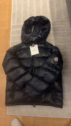 Moncler Maya - Säljer en mörkblå, glansig pufferjacka från Moncler med huva och dragkedja. Jackan har Moncler-logga på ärmen och två fickor med dragkedja. Perfekt för kalla dagar och riktigt snygg streetstil. Bvsa. Intyg fås såklart och vidarebefordras via mail.