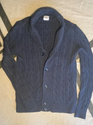Kabelstickd kofta från John Henric - Säljer en mörkblå kabelstickad kofta från John Henric i storlek S. Koftan har snygg sjalkrage, fyra knappar framtill och ribbade muddar vid ärmslut och nederkant. Perfekt för lager-på-lager och chill stil under höst och vinter.