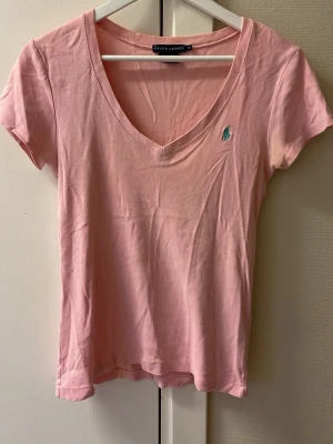 Rosa v-ringad t-shirt från Ralph Lauren - Säljer en ljusrosa t-shirt från Ralph Lauren med v-ringning och korta ärmar. T-shirten har en broderad logga i turkos på bröstet och är gjord i mjuk bomull. Perfekt för dig som gillar klassisk stil med en twist.
