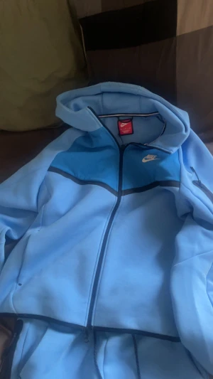 Nike ljusblå mjukisbyxor med snörning - Nike mjukisbyxor i ljusblått med mörkare blå paneler på knäna och svarta detaljer. Byxorna har snörning i midjan och dragkedja på ena benet. Perfekta för chill eller träning, med klassisk Nike-logga på benet och tröjan ingår helt nya använt typ 2-3 gånger säljer dom för dom inte passar 