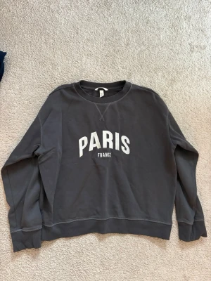 Mörkgrå sweatshirt med Paris-tryck - Säljer en mörkgrå sweatshirt från H&M med vitt Paris France-tryck på bröstet. Tröjan har rund hals, långa ärmar och ribbade muddar vid ärmslut och nederkant. Perfekt för chill dagar och enkel att matcha med jeans eller joggers.