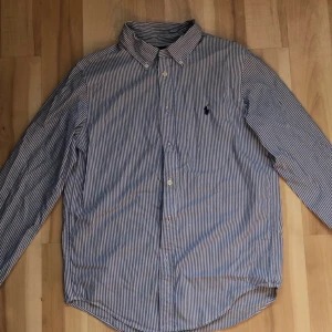 Blårandig skjorta från Ralph Lauren - Snygg blårandig skjorta från Ralph Lauren med klassisk krage och broderad logga på bröstet. Skjortan har lång ärm, knappar framtill och är gjord i mjuk bomull. Passformen är custom fit vilket ger en schysst siluett. Perfekt för dig som gillar stilrena och tidlösa plagg. Storlek s