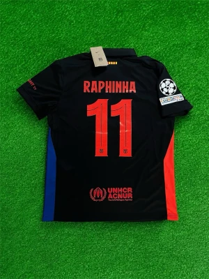 Raphinha fotbollströja Barcelona  - Bilderna visar en fotbollströja från FC Barcelona med Raphinha och nummer 11, tillverkad av Nike. Tröjan är svart med röda och blå detaljer, Champions League-märke på ärmen och UNHCR-logga. Ingen sko syns på bilderna.