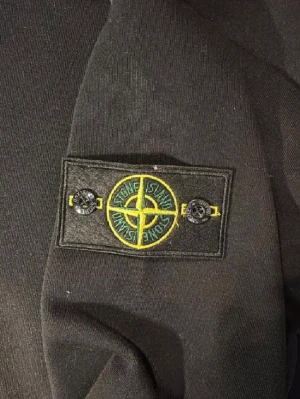 Svart sweatshirt från Stone Island - Svart långärmad sweatshirt från Stone Island med klassisk rund hals och ikonisk patch med logga på vänster ärm. Tröjan har en clean look och är tillverkad i mjuk bomull som känns skön mot huden. Perfekt för dig som gillar streetwear och stilrena plagg.
