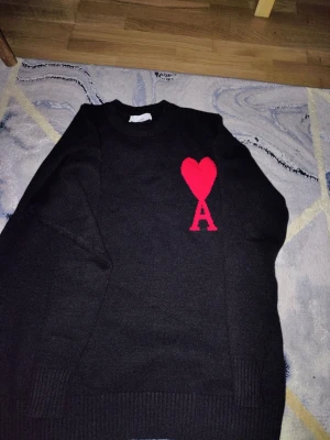 Svart stickad tröja med rött hjärta - Svart stickad tröja med rund hals och ett stort rött hjärta samt bokstaven A på bröstet. Tröjan har långa ärmar och ribbade muddar vid ärmslut och nederkant. Perfekt för dig som gillar stilrena plagg med en twist.
