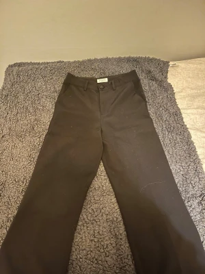 Svarta kostymbyxor från Lindex - Svarta kostymbyxor från Lindex med klassisk rak passform och midja med hällor för bälte. Byxorna har diskreta fickor bak och fram, samt knapp och dragkedja i midjan. Perfekta för en clean och stilren look.