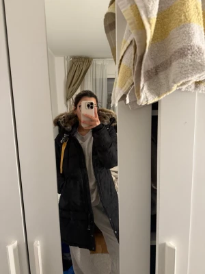 Svart Parajumpers dunjacka med päls - Säljer en svart lång dunjacka från Parajumpers med stor huva och pälsdetalj. Jackan har en gul rem vid kragen, dragkedja framtill och flera fickor. Perfekt för kalla dagar och riktigt snygg streetstil. Passar xs, S och M