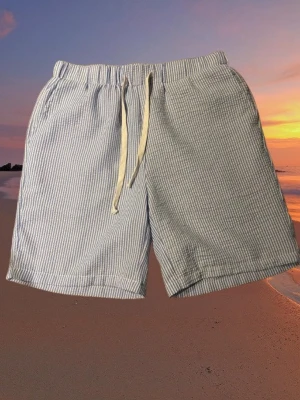 Randiga linne shorts med snörning - Säljer ett par ljusblå och vita randiga shorts med elastisk midja och snörning framtill. Shortsen har sidofickor och en bakficka, perfekta för sommardagar på stranden. Tillverkade i ett lätt och svalt linne och bomullstyg.